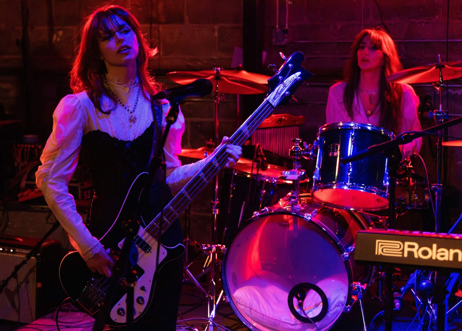 Mercedes on bass at the mic with Mónica behind the kit, lit red and blue