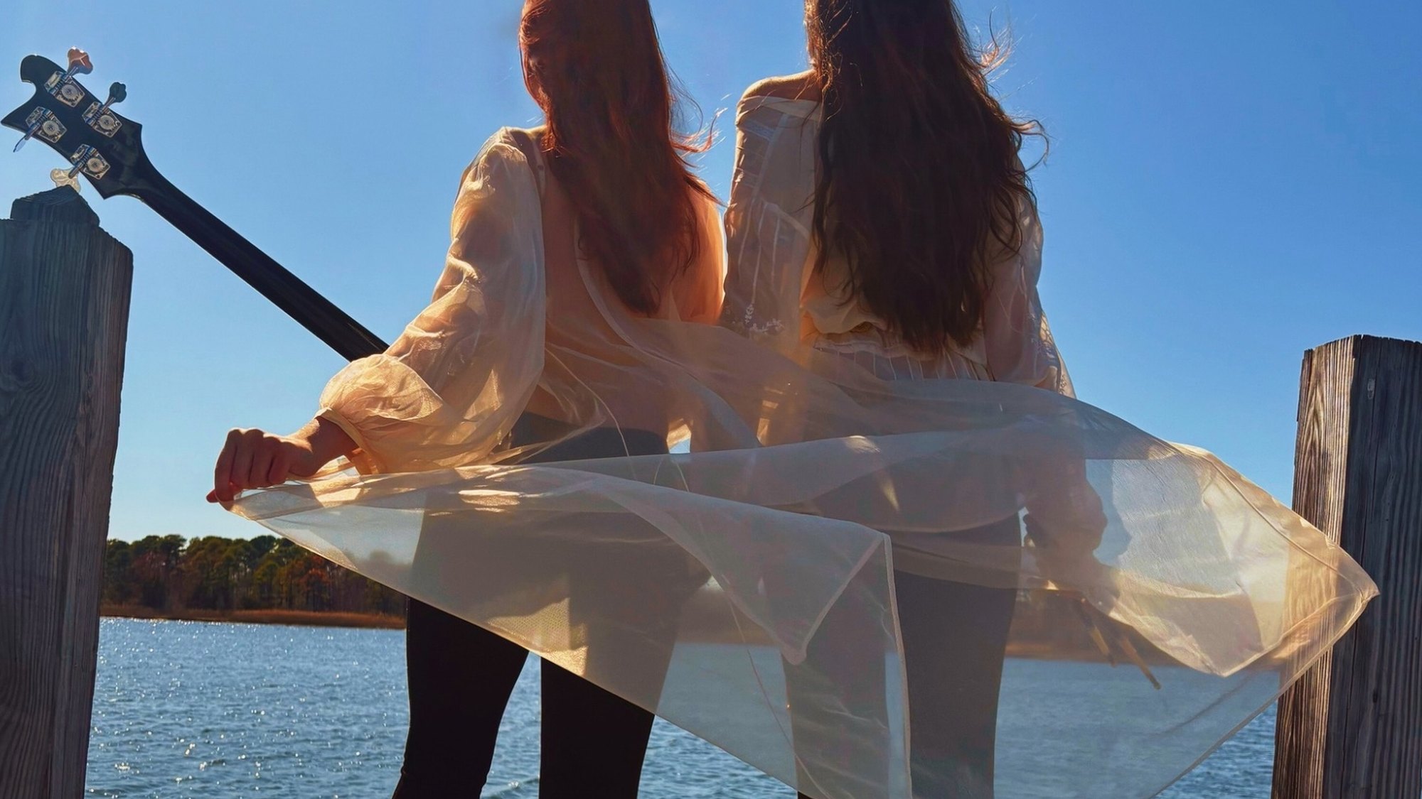 Mónica and Mercedes of Söūp standing at the end of a dock over the water, a bass slung on one shoulder